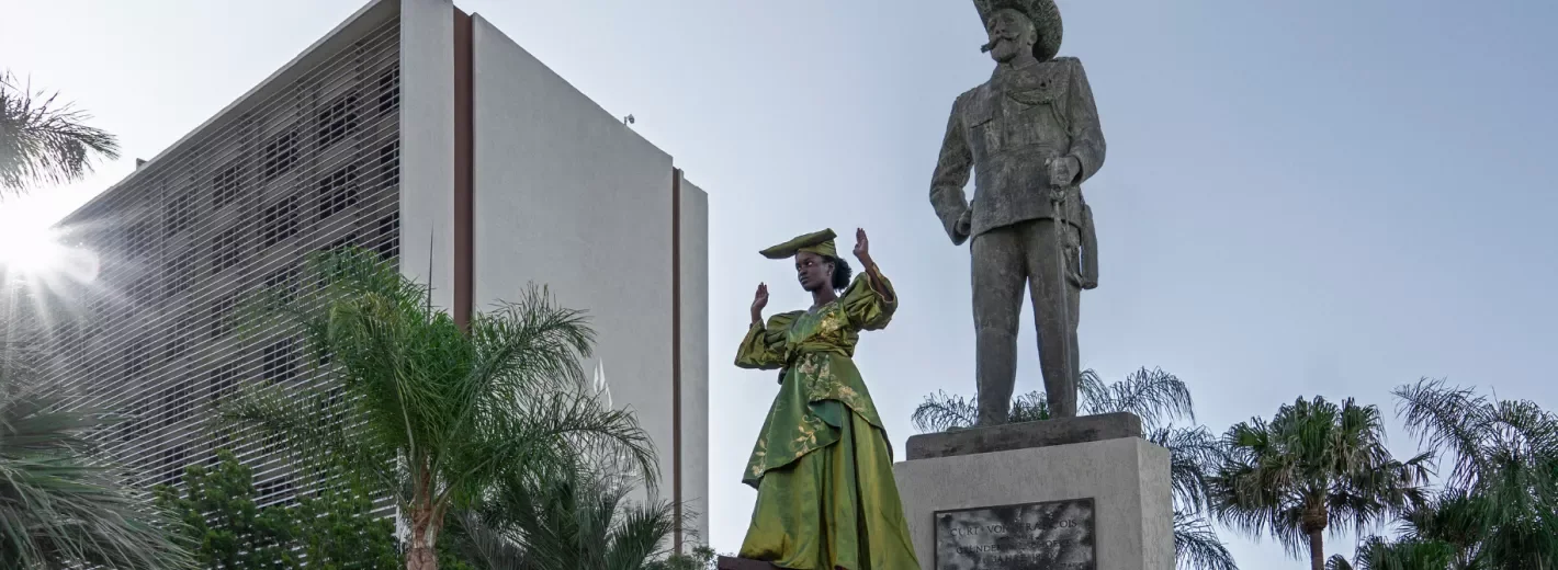 Outdoor scene showing a person wearing a bright green costume performing with raised arms beside a large bronze statue on a stone pedestal, set among palm trees and modern buildings in a sunlit urban environment.