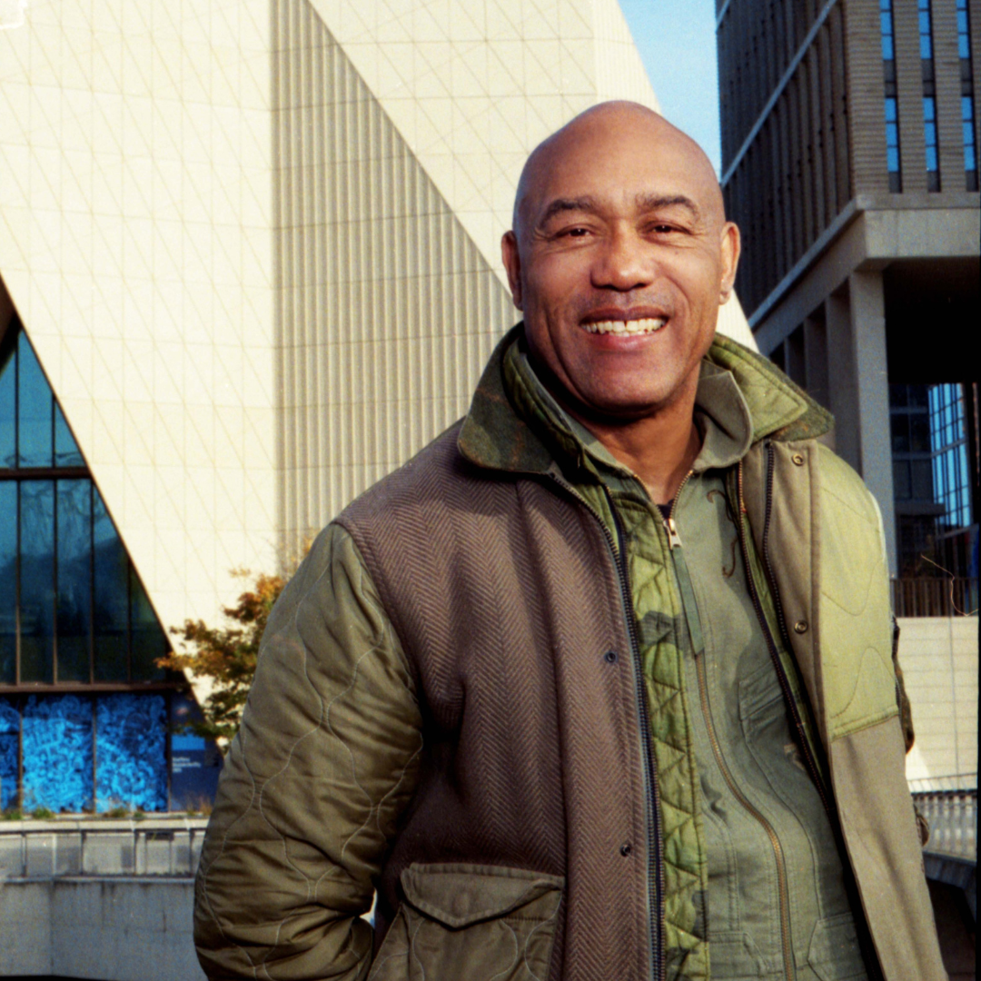 Person standing outdoors in front of the V&A Dundee building, with the museum’s angular, modern architecture visible behind them. The building features distinctive geometric forms, textured concrete surfaces and large glass sections.