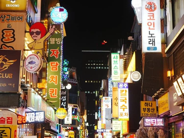 Nighttime street scene with bright neon signs in Korean lining both sides of a narrow, busy street, featuring illuminated shopfronts, vertical signboards, and dense urban buildings glowing against a dark sky.