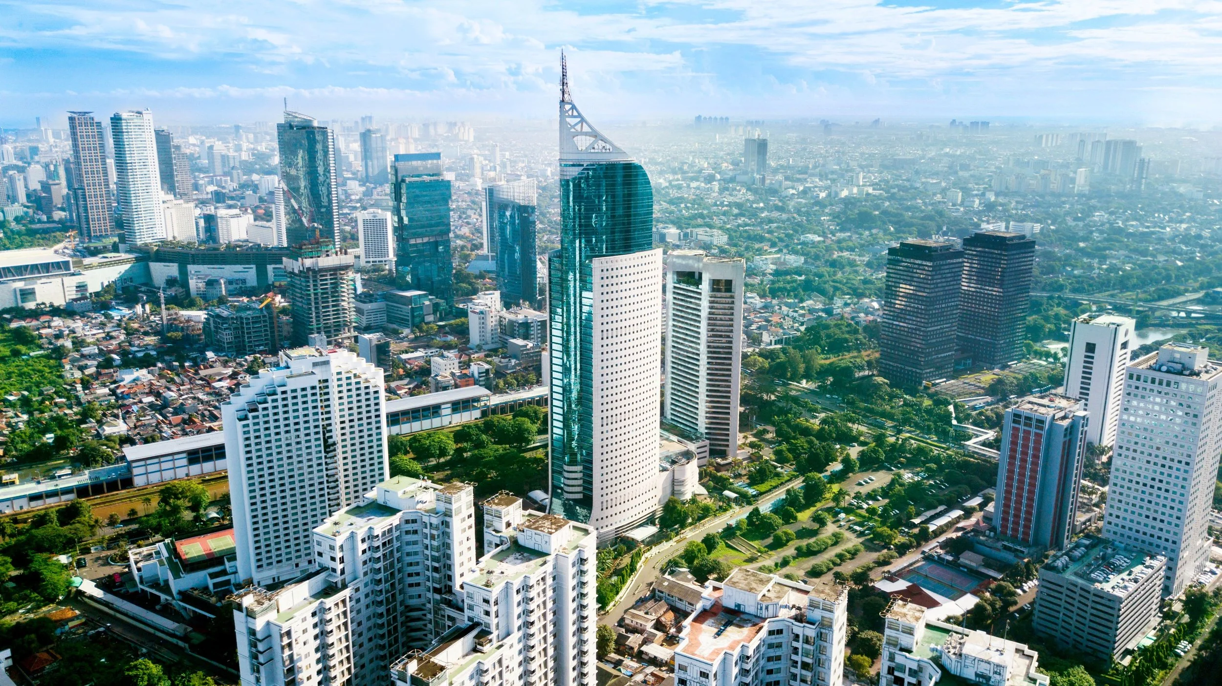 Aerial view of a dense city with modern high‑rise buildings, office towers and residential blocks surrounded by tree‑lined roads, with a hazy skyline stretching into the distance under a bright blue sky.