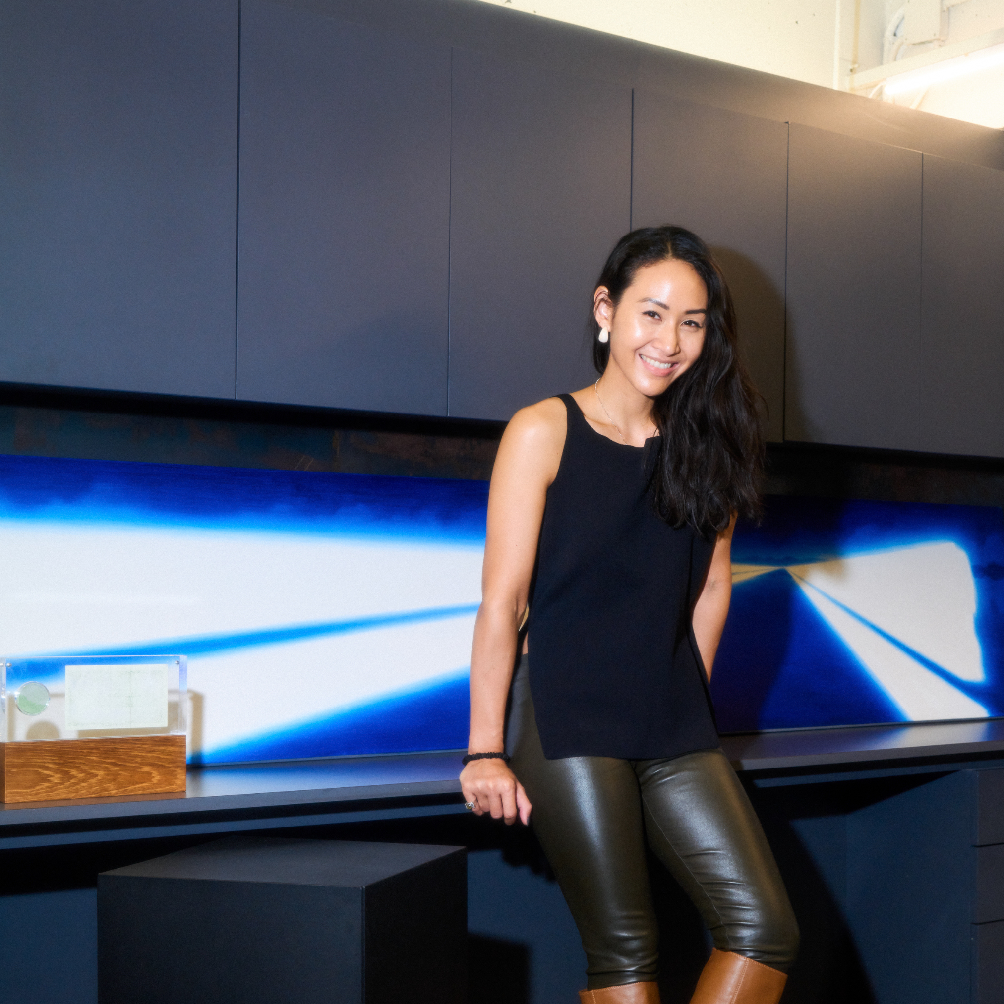 Person standing indoors in a contemporary exhibition or gallery space, leaning against a black cabinet beneath a long, horizontal illuminated artwork displaying abstract blue and white light patterns.
