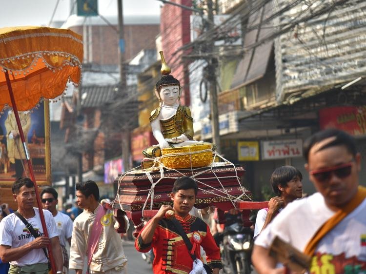 Street procession carrying a seated Buddha statue on a decorated platform, held aloft by participants walking through a busy urban street lined with buildings, signage and overhead cables, with onlookers gathered around.