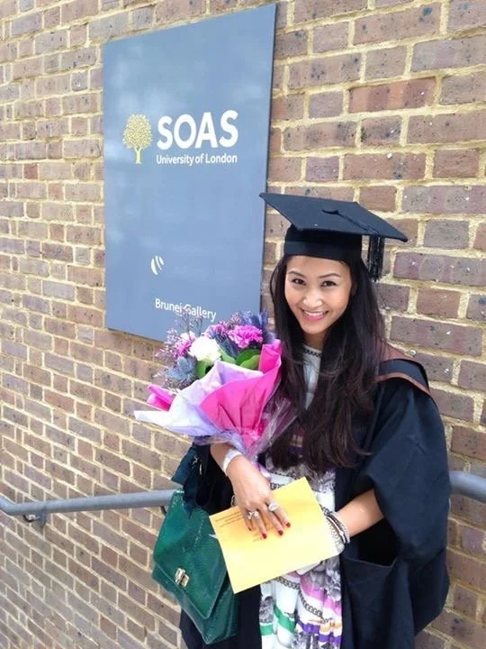 Graduate wearing academic gown and mortarboard standing outside the Brunei Gallery at SOAS University of London, holding a bouquet of flowers and an envelope, with a blue SOAS sign mounted on a brick wall behind them.