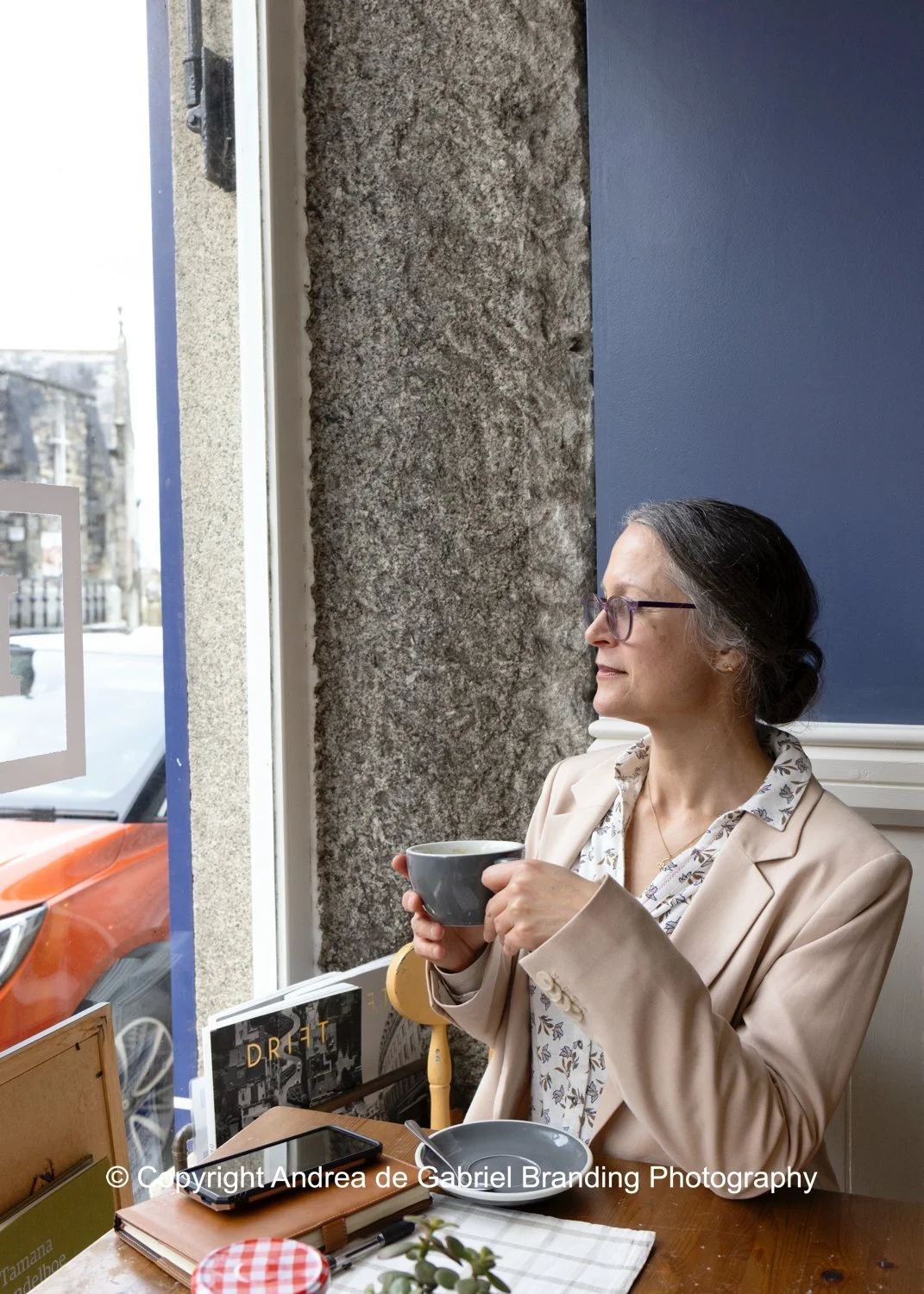 A woman with gray hair and glasses sitting in a cafe, holding a cup of coffee, looking out the window.