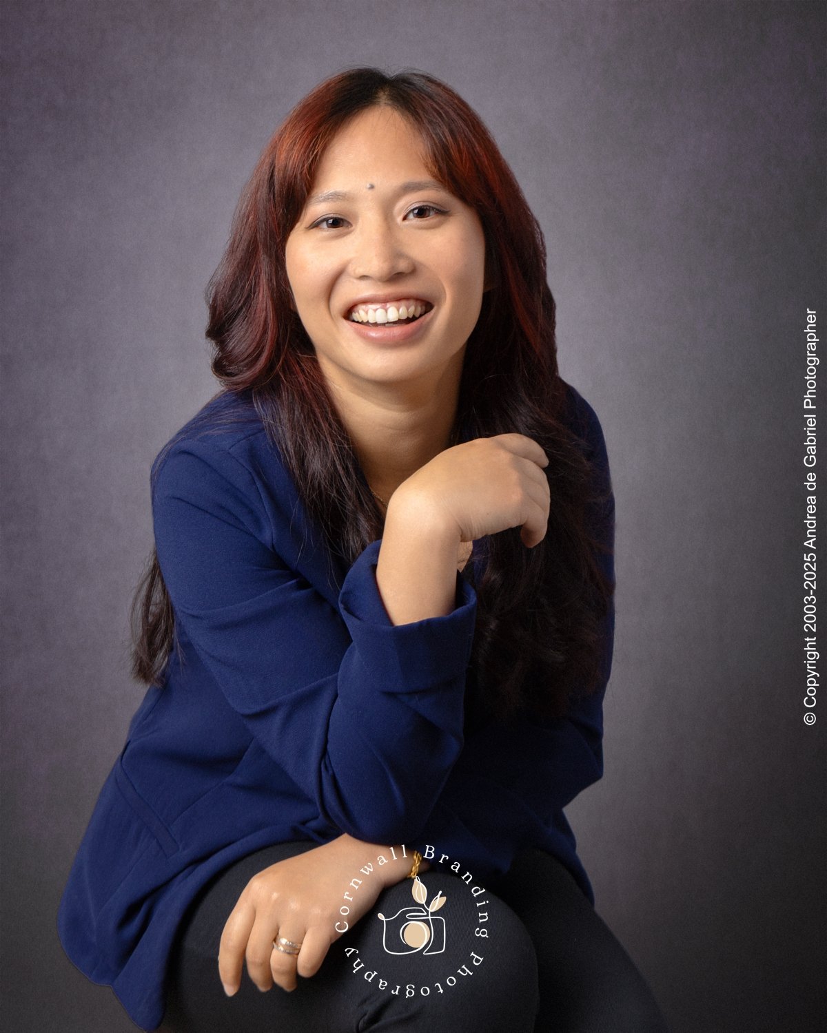 Woman with long dark hair smiling, wearing a dark blue blazer, sitting against a gray background.