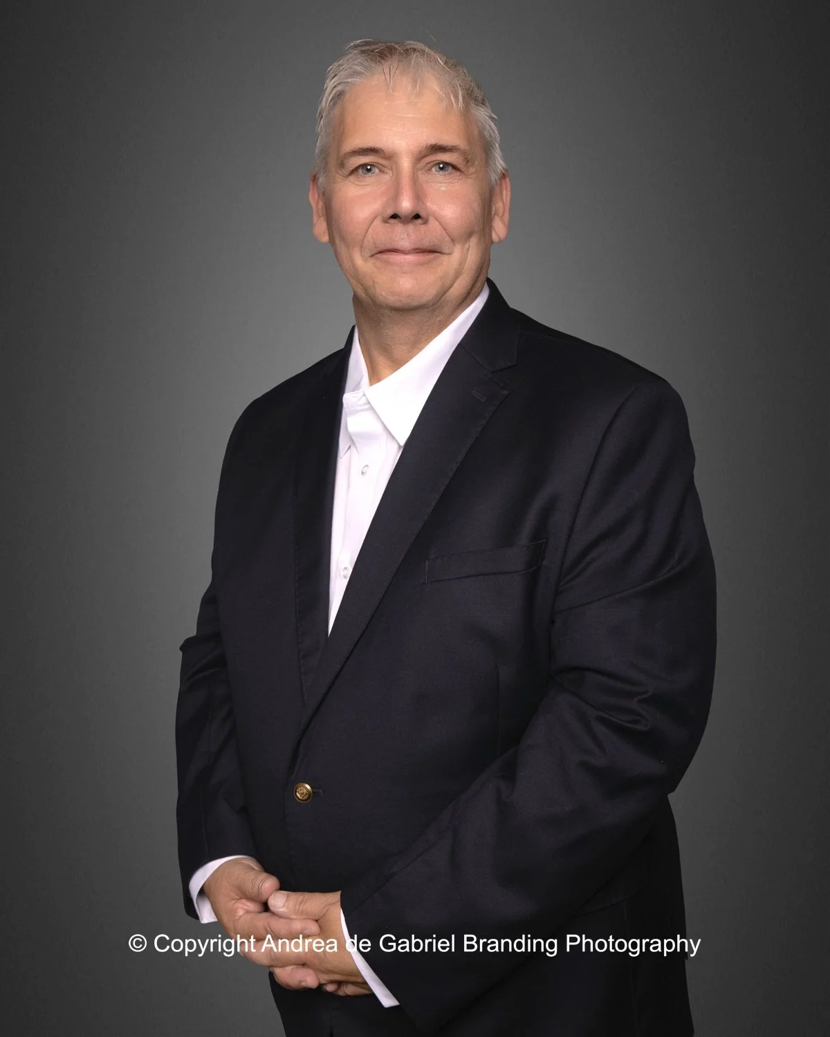 A middle-aged man with gray hair, wearing a black suit and white shirt, stands against a gray background, smiling gently.
