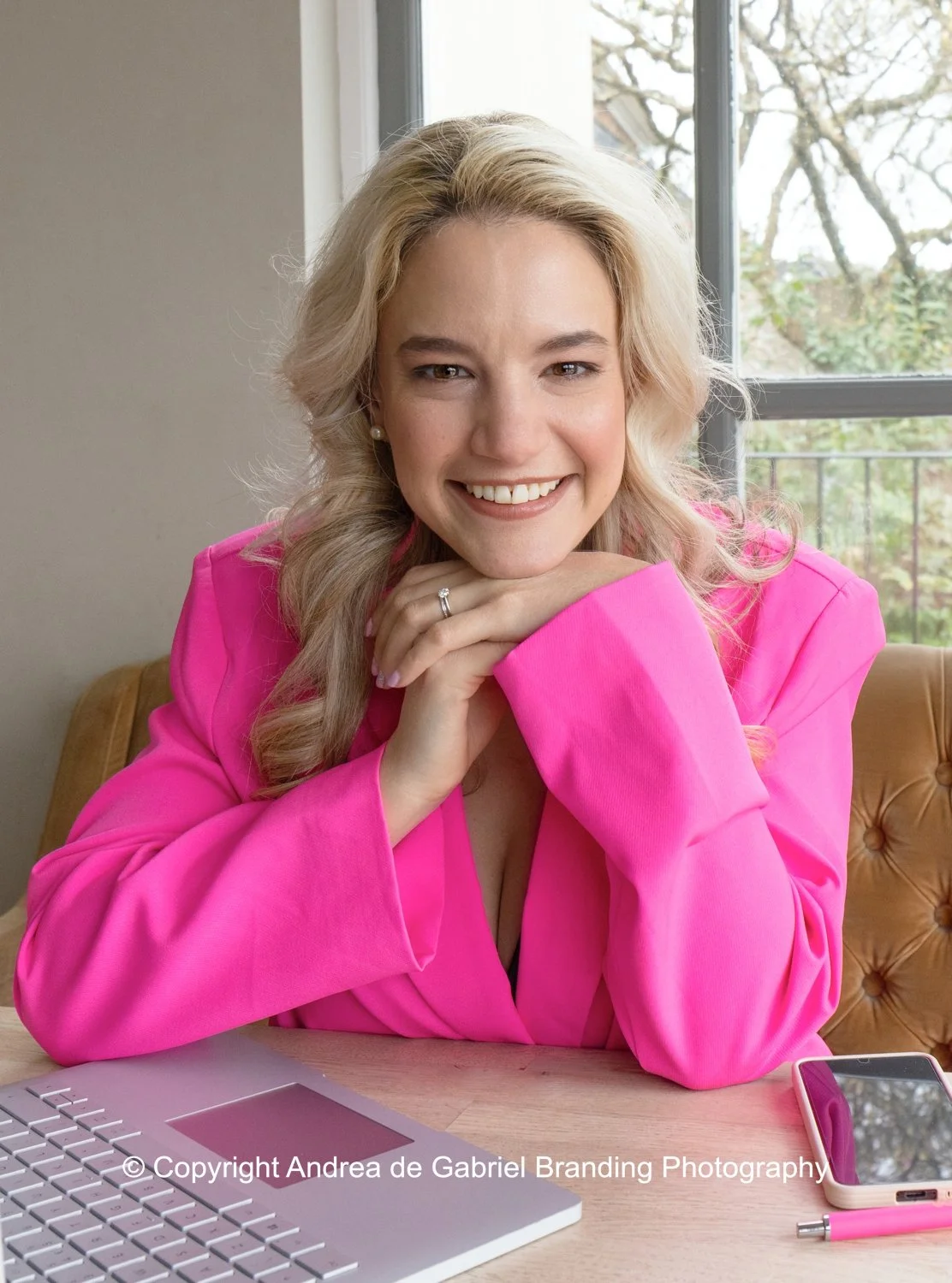 A smiling woman with blonde hair in a pink blazer sitting at a desk with a laptop and smartphone by a window.