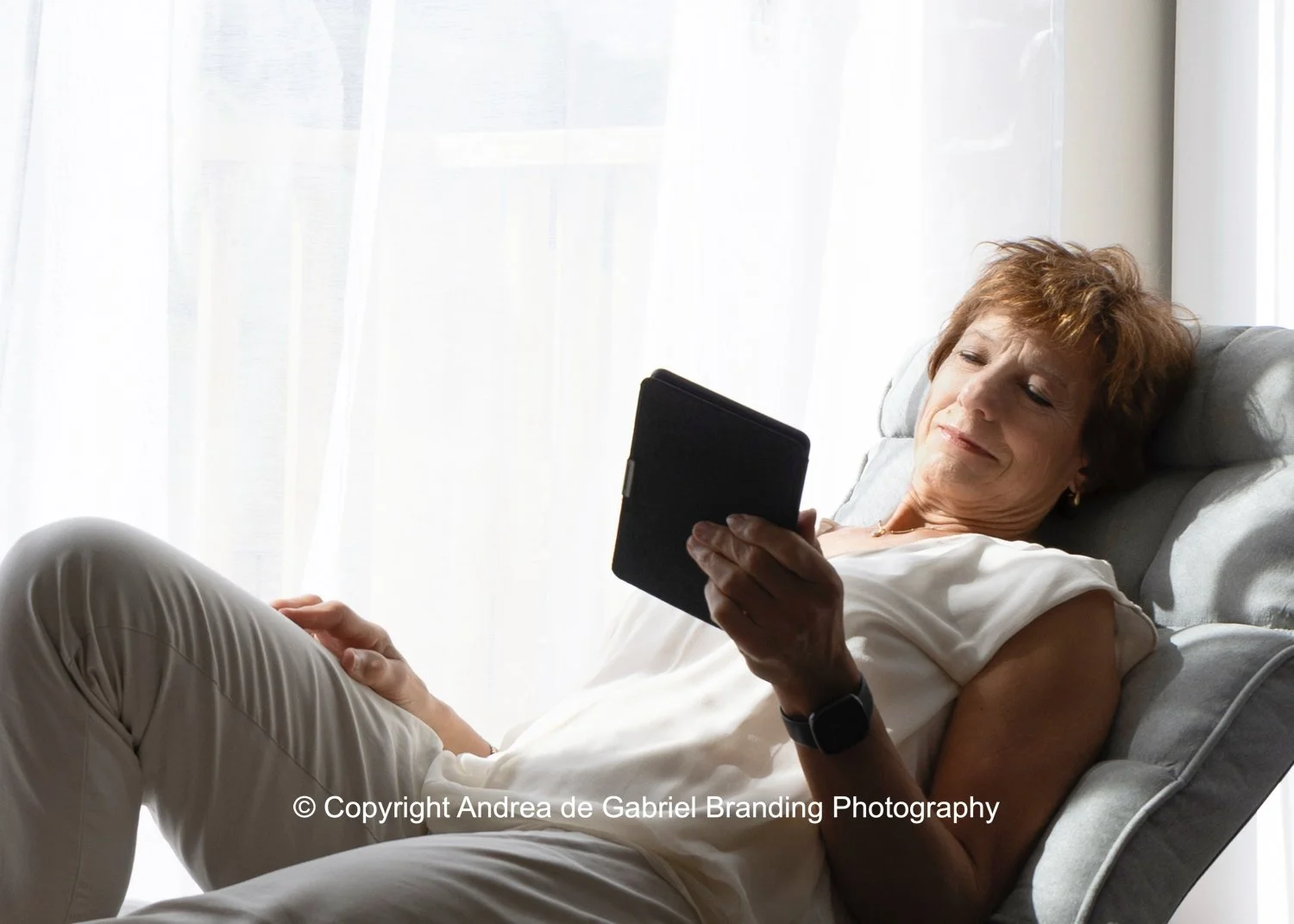 An elderly woman with short red hair reclining on a gray chair, reading a black tablet, illuminated by natural light from a nearby window.