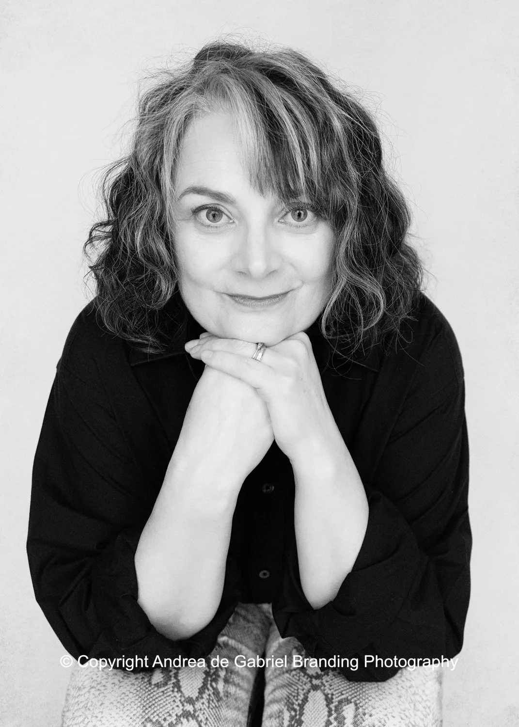 Black and white portrait of a woman with curly hair, smiling gently, resting her chin on her clasped hands, wearing a dark blouse.