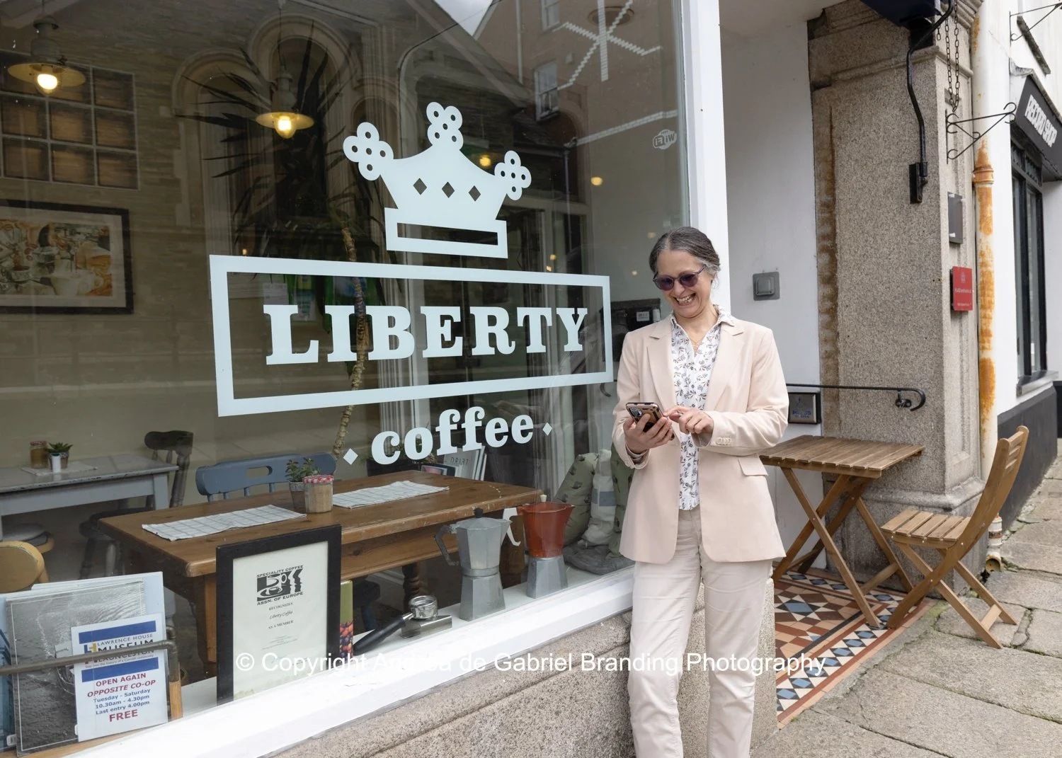 Woman in beige suit looking at her phone outside a coffee shop named Liberty Coffee, with large glass window decorated with the shop's logo, some tables, and chairs on the sidewalk.