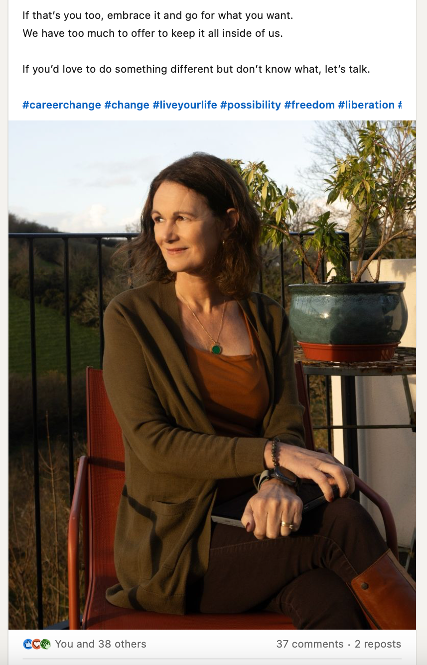 A woman with shoulder-length hair sits outdoors on a red chair, thoughtfully resting their head on their hand. They wear a dark green top and brown pants, surrounded by potted plants under a clear sky.