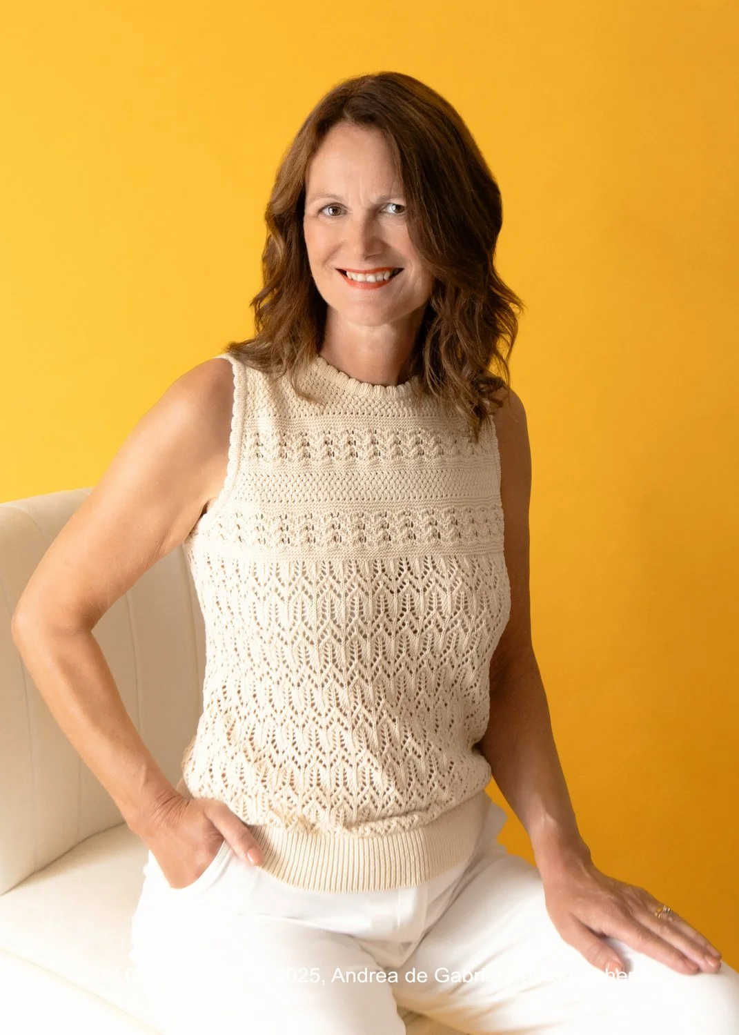 A woman with shoulder-length brown hair in a sleeveless, cream-colored knitted top and white pants, standing against a yellow background, smiling with her right hand in her pocket and her left hand resting on a white surface.