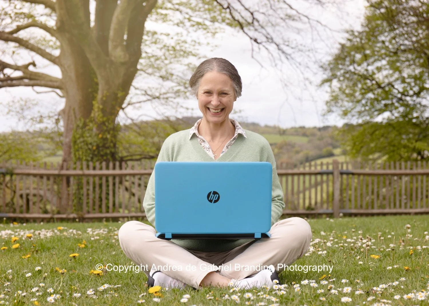 A woman sitting cross-legged on grass in a park, using a light blue HP laptop, smiling, with trees and a fence in the background.
