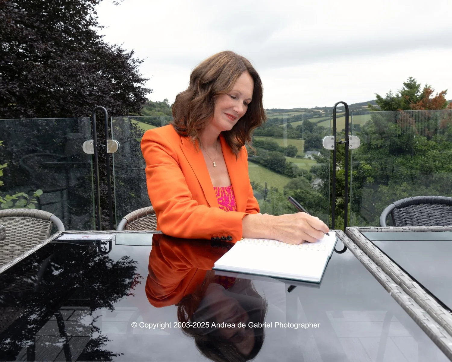 A woman in an orange blazer sitting at an outdoor table, writing in a notebook, with a scenic green landscape in the background.