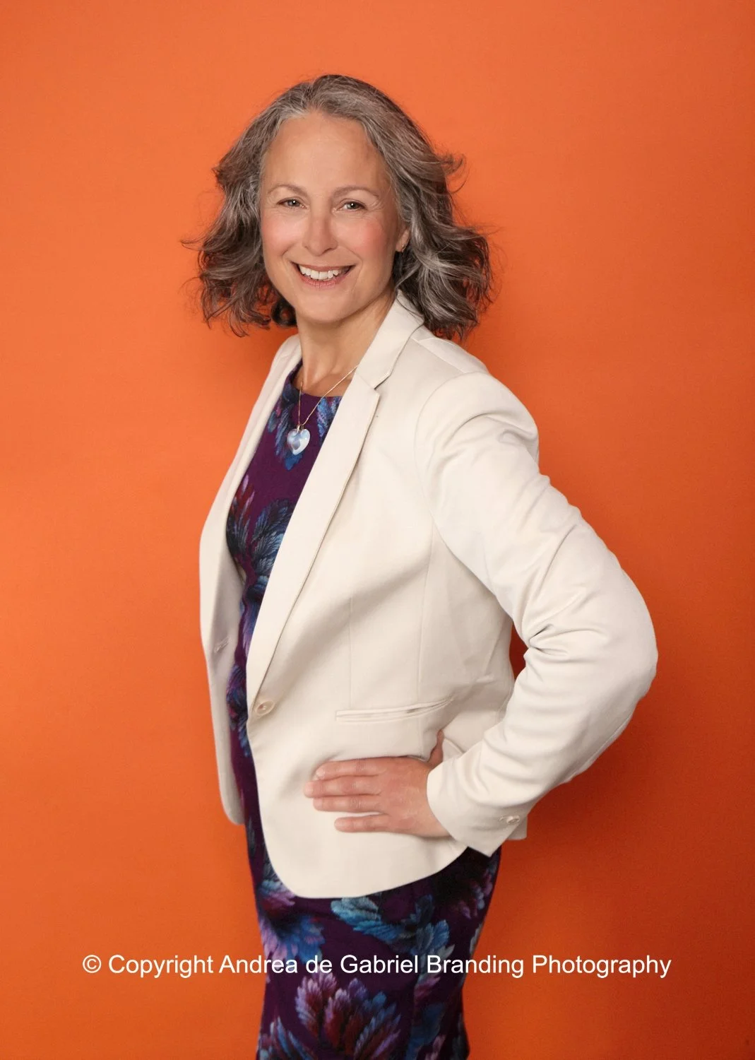 A woman with shoulder-length gray hair, wearing a white blazer over a colorful dress, smiling with one hand on her hip against an orange background.