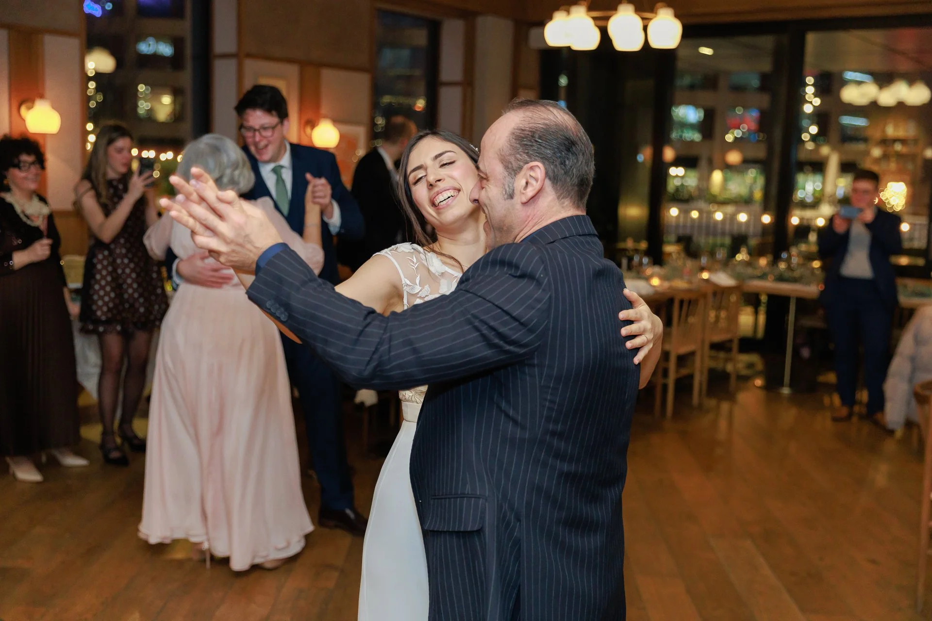 A woman and an older man are dancing closely at a celebration, smiling happily, surrounded by other guests in a warmly lit venue.