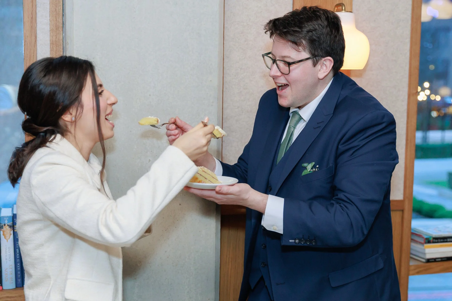 A man and woman sharing a piece of cake, with the man holding a plate of cake for the woman, both smiling, indoor setting