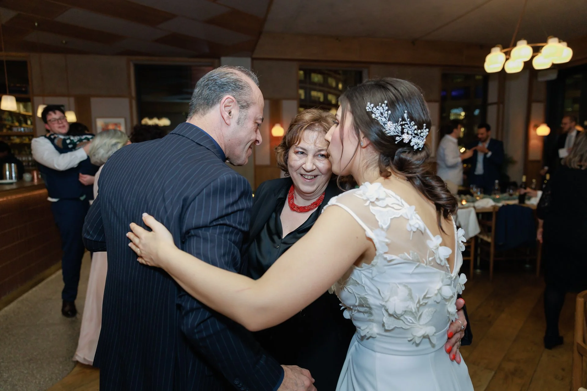 A bride in a white wedding dress with lace and floral applique is kissing a man in a dark suit, while a woman in black holds them and smiles at the camera. They are at a wedding reception with other guests in the background.
