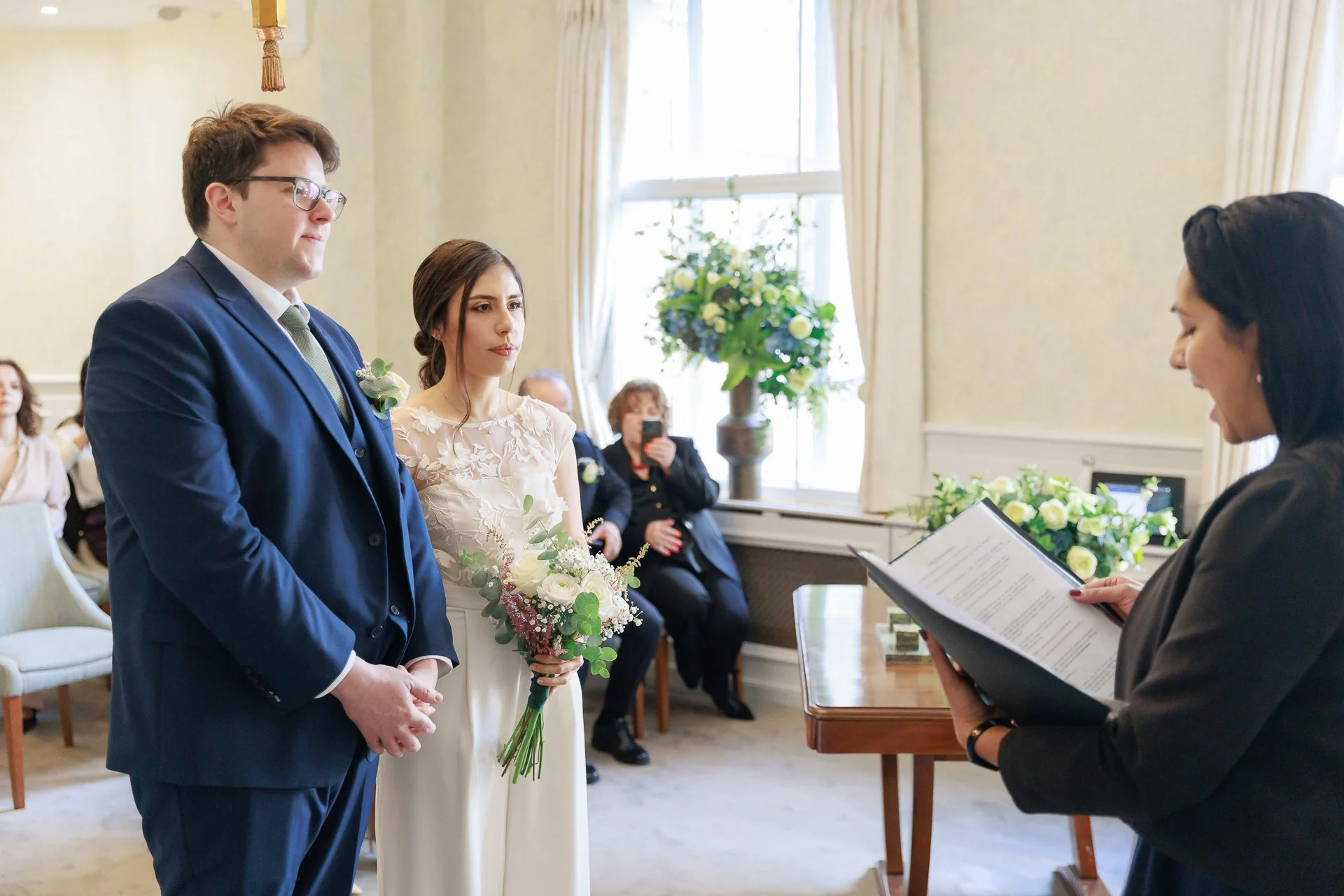 A couple stands at the altar during a wedding ceremony, with the officiant reading from a book. The groom is wearing a dark blue suit with a white shirt and tie, and glasses. The bride is dressed in an elegant white gown, holding a bouquet of white a