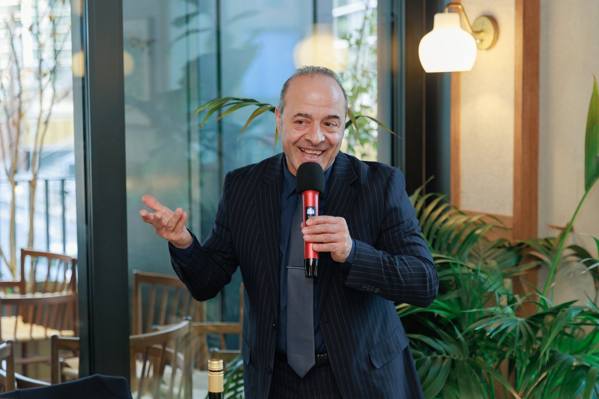 A man in a business suit holding a microphone, gesturing with his hand, and smiling while giving a speech or presentation indoors.