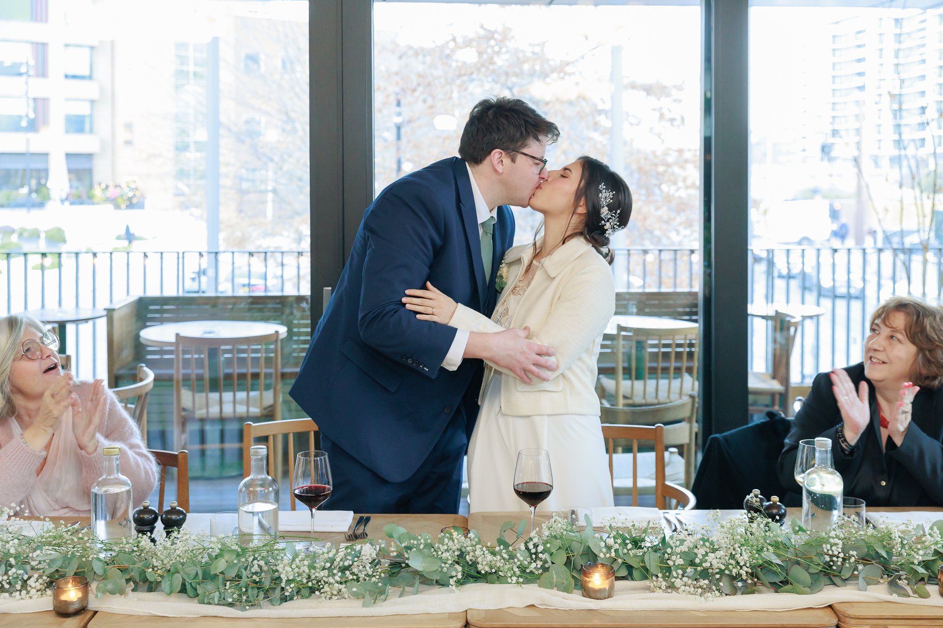 A newlywed couple sharing a kiss during a wedding reception, surrounded by family members who are clapping and celebrating at a decorated banquet table with flowers and candles.