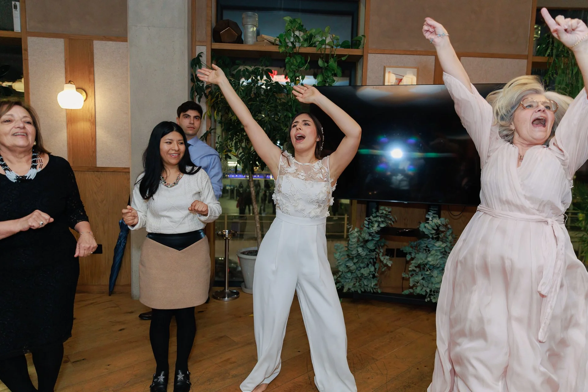 Group of women and a man dancing and singing at an indoor celebration with plants and wooden decor.