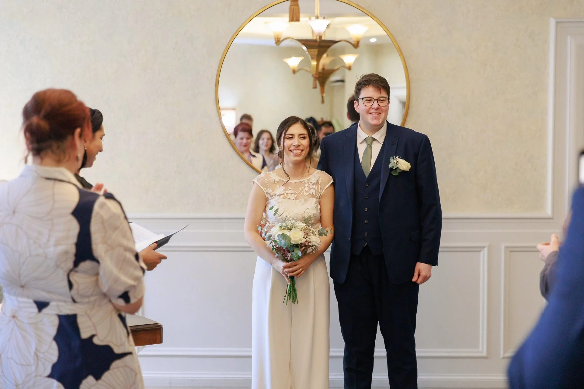 A wedding ceremony with a bride and groom standing together, holding a bouquet of flowers, smiling in front of a mirror reflecting guests, with officiants and guests witnessing.