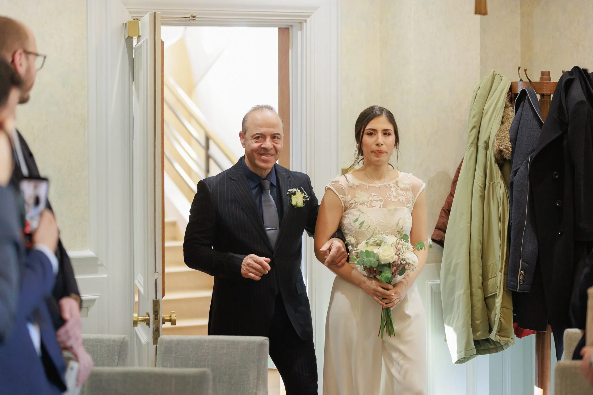 A bride walking down the aisle accompanied by an older man, possibly her father, in a wedding ceremony. The bride is holding a bouquet of flowers and has a neutral expression. The setting appears to be indoors, with guests visible on the sides.