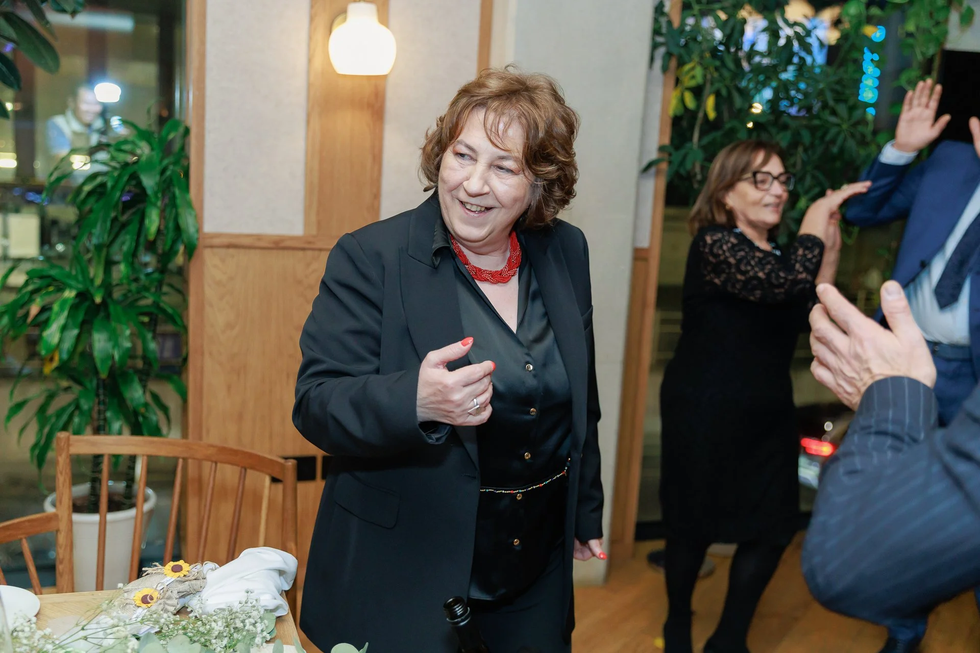 A woman with curly brown hair wearing a black blazer and red necklace smiling while talking to someone at a social gathering. In the background, another woman with glasses and black dress is talking and gesturing. The setting appears to be a restaura