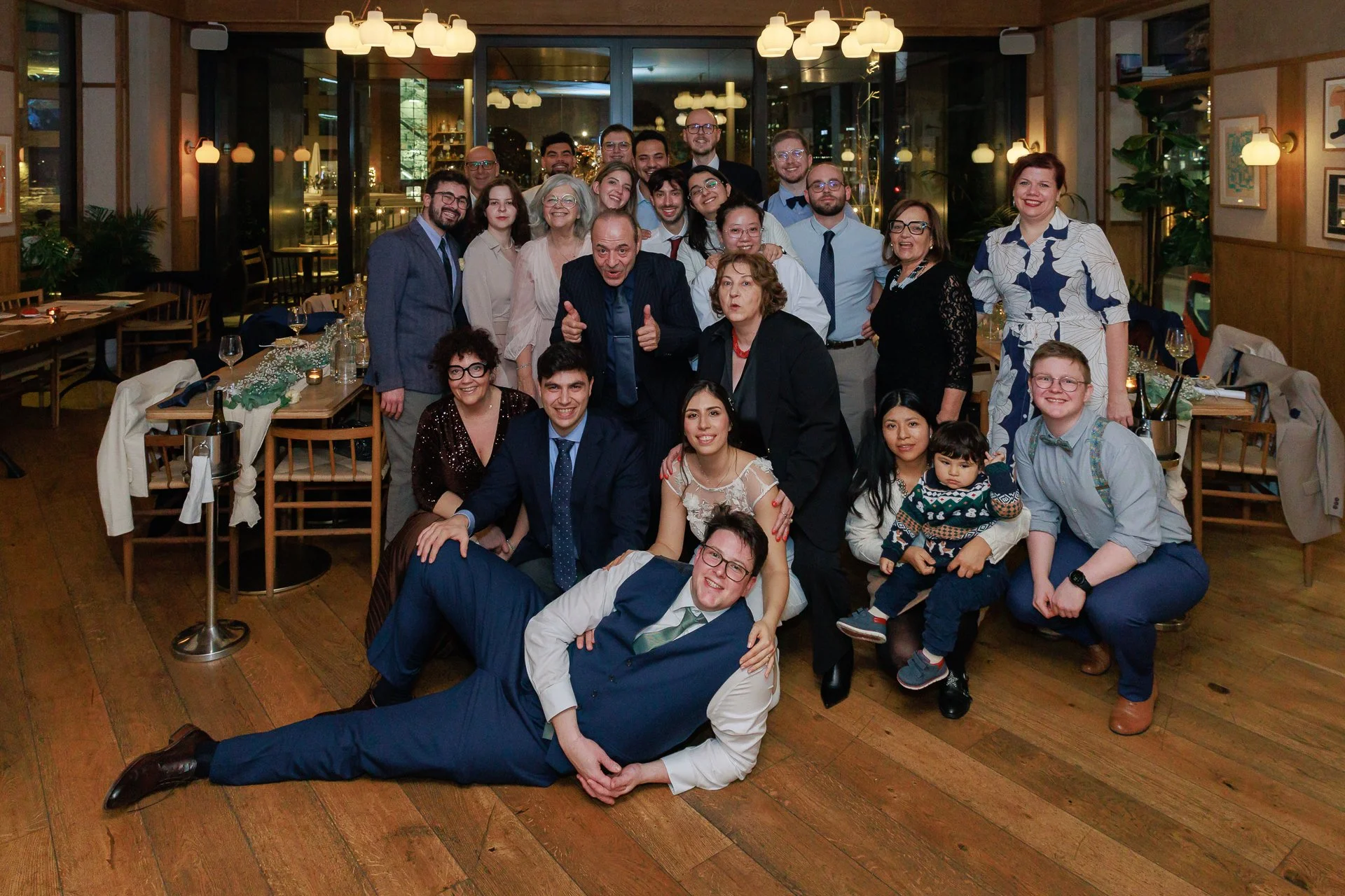 A group of people celebrating at a restaurant, posing for a photo with some sitting and others standing behind. The setting has warm lighting, wooden furniture, and decorated tables.
