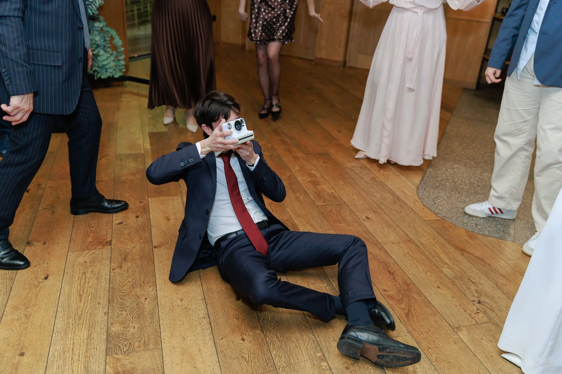 Man in a suit and red tie sitting on the wooden floor at a formal event, taking photos with a vintage camera, surrounded by other formally dressed people.