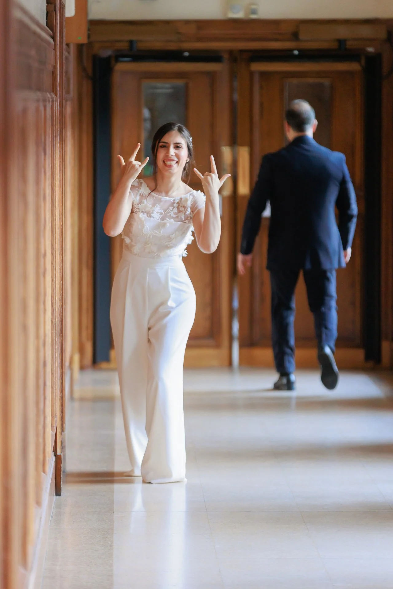 Woman in white pants and lace top smiling and making a rock and roll hand gesture in a wooden hallway, man in dark suit walking away in the background.