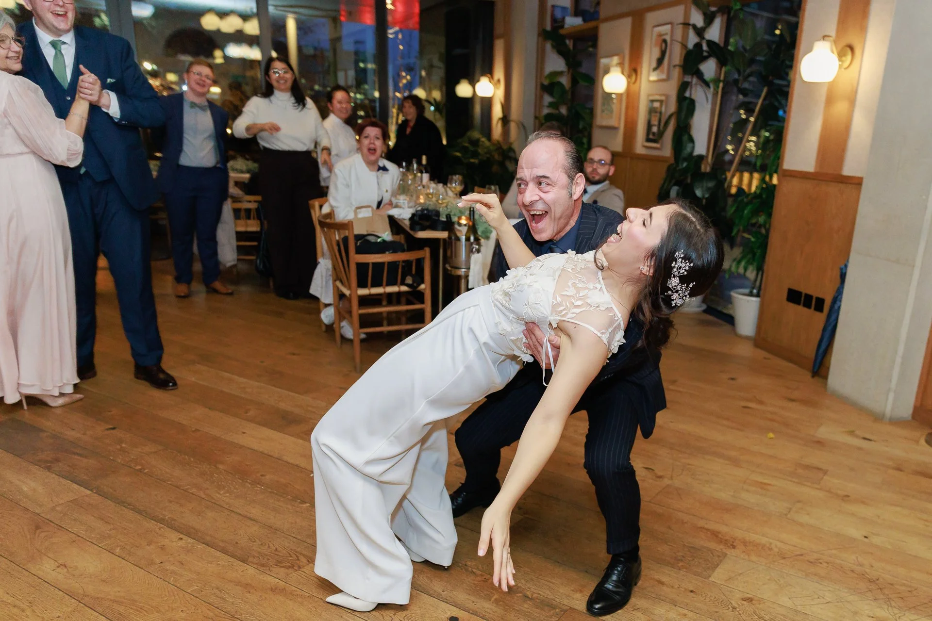 A man and woman dancing playfully at a celebration, with the man dipping the woman back as she laughs, surrounded by seated and standing guests in a warmly lit restaurant setting.