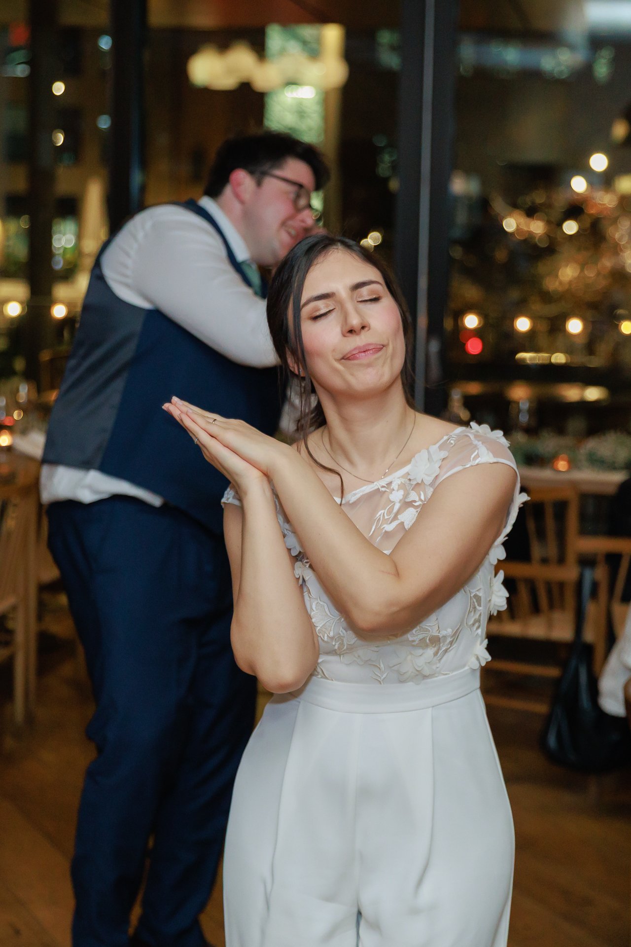 A woman in a white dress with floral embroidery making a playful, pouting expression, with her eyes closed and lips slightly pursed. A man in a vest and glasses is in the background, leaning on the woman’s head with a smile. The setting appears to be