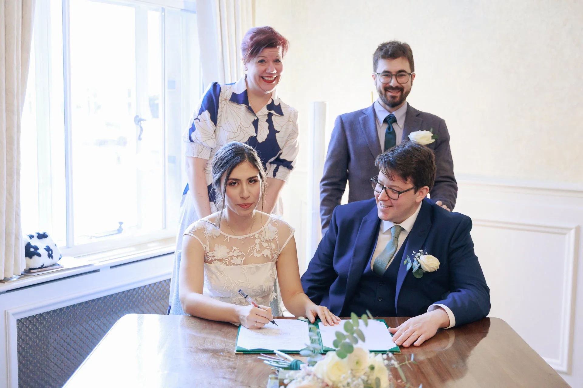 A bride and groom sign their wedding register while two guests look on smiling indoors near a window with curtains.