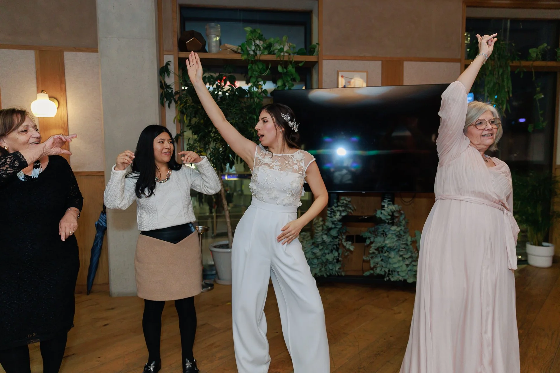 Four women dancing and enjoying themselves at a celebration. The woman in the center is wearing a white dress with lace details and high-waisted white trousers with her right hand in the air. The woman on the far right is dressed in a pink dress with