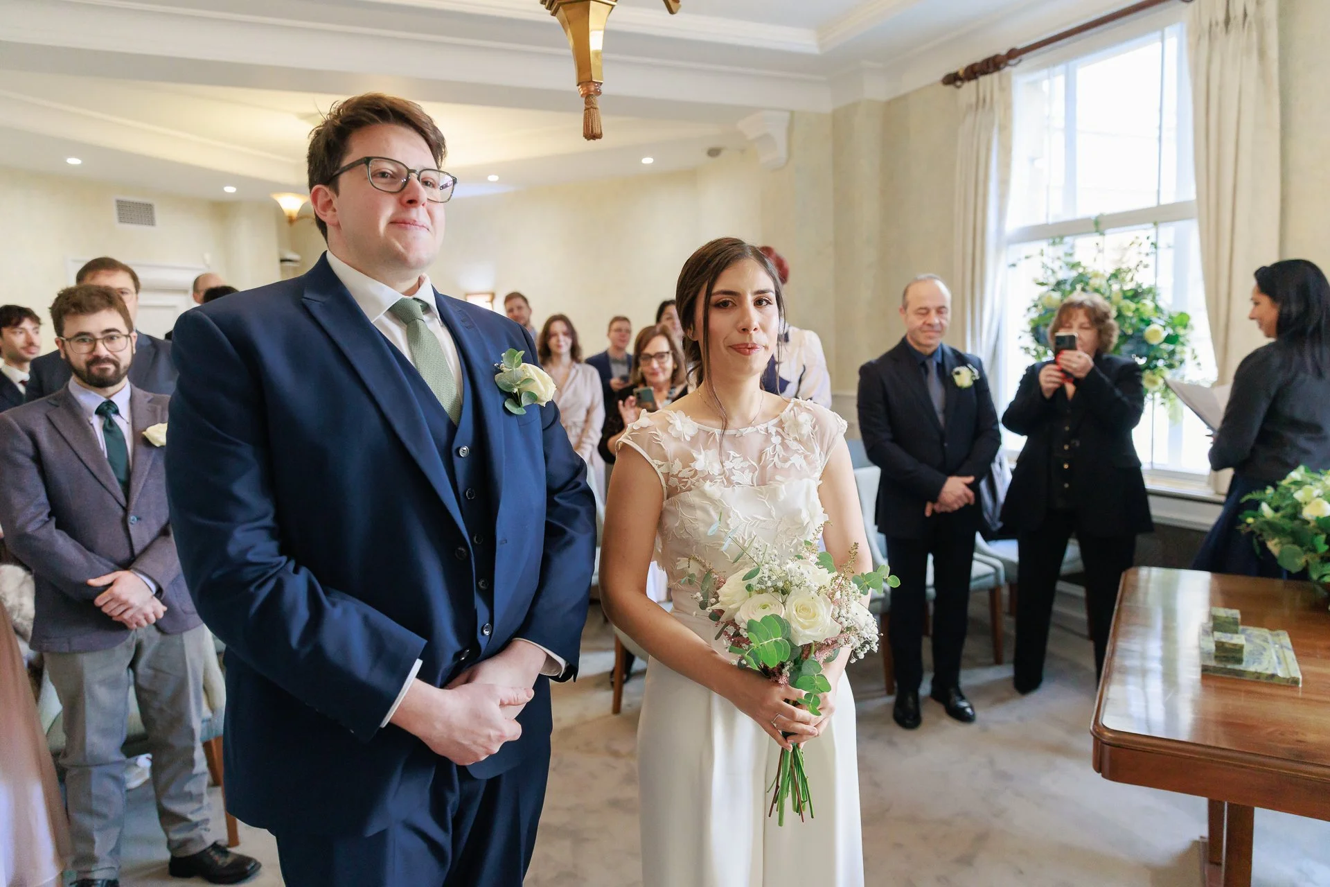 A wedding ceremony with a bride holding a bouquet and a groom in a blue suit, surrounded by guests in a decorated room with windows and curtains.