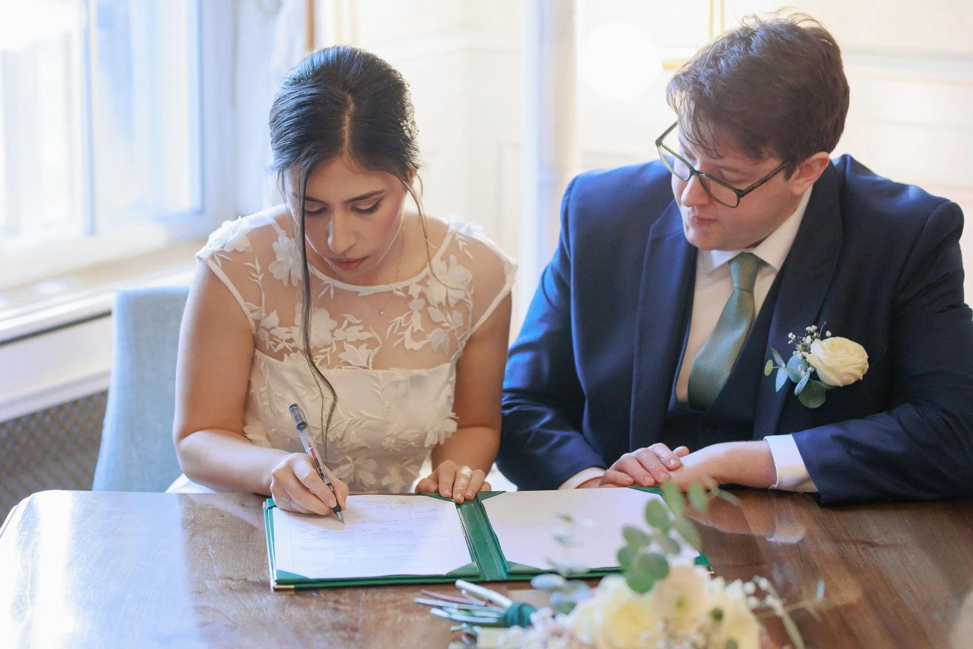 A woman and a man sitting at a table, signing a document during a wedding ceremony. The woman is wearing a white dress with floral lace details, and the man is dressed in a navy suit with a white shirt, green tie, and a white flower boutonniere.