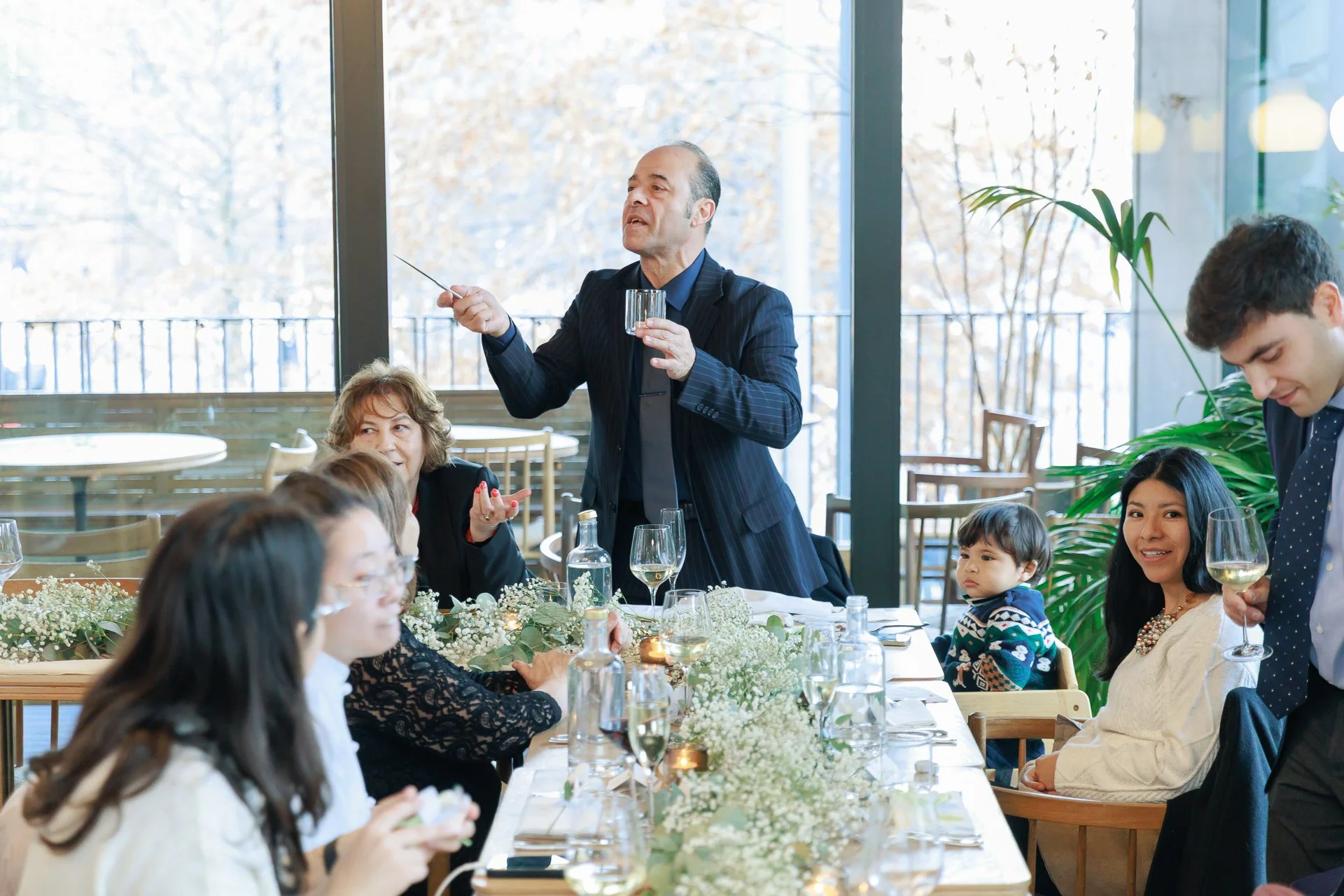 Man giving speech at a family gathering, with women and children sitting at a decorated table with wine glasses and bottles.