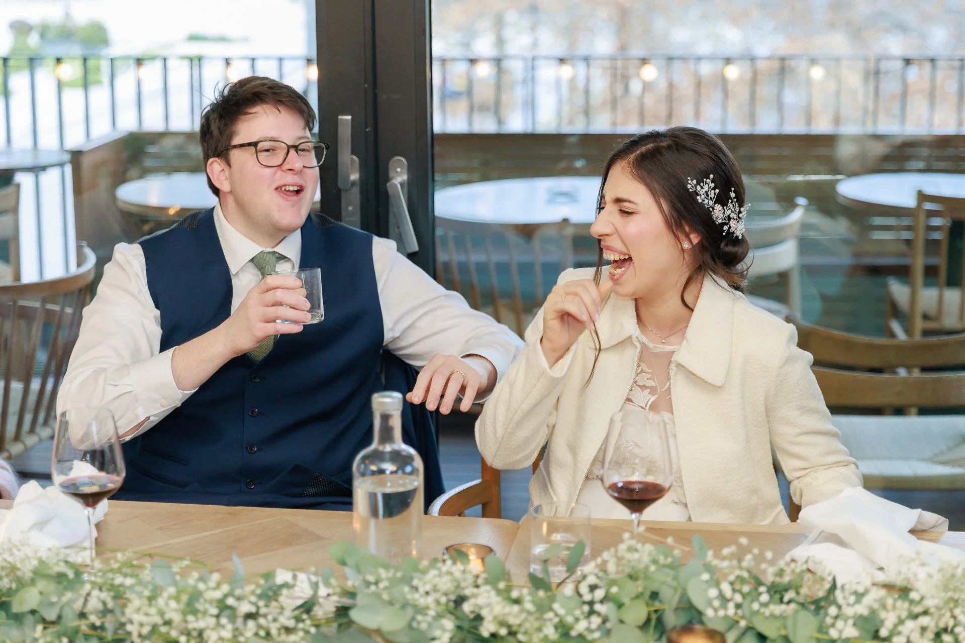 A man and a woman laughing and enjoying drinks at a wedding reception table, with wedding decorations and a view of a patio outside.