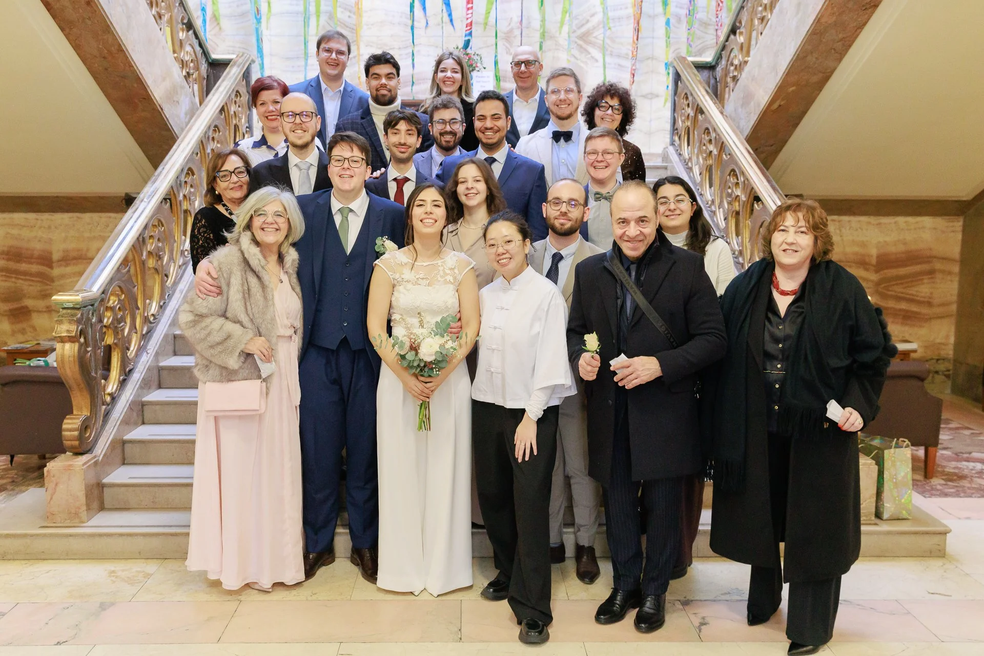 Group of wedding guests posing on a staircase, with the bride and groom in the center holding a bouquet of flowers, all smiling.