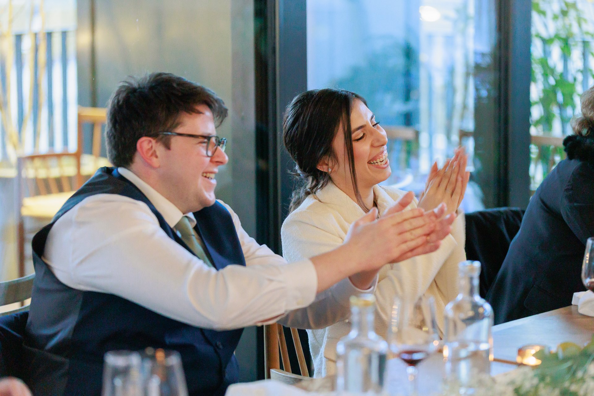 People laughing and clapping at a dinner event with wine bottles and glasses on the table.
