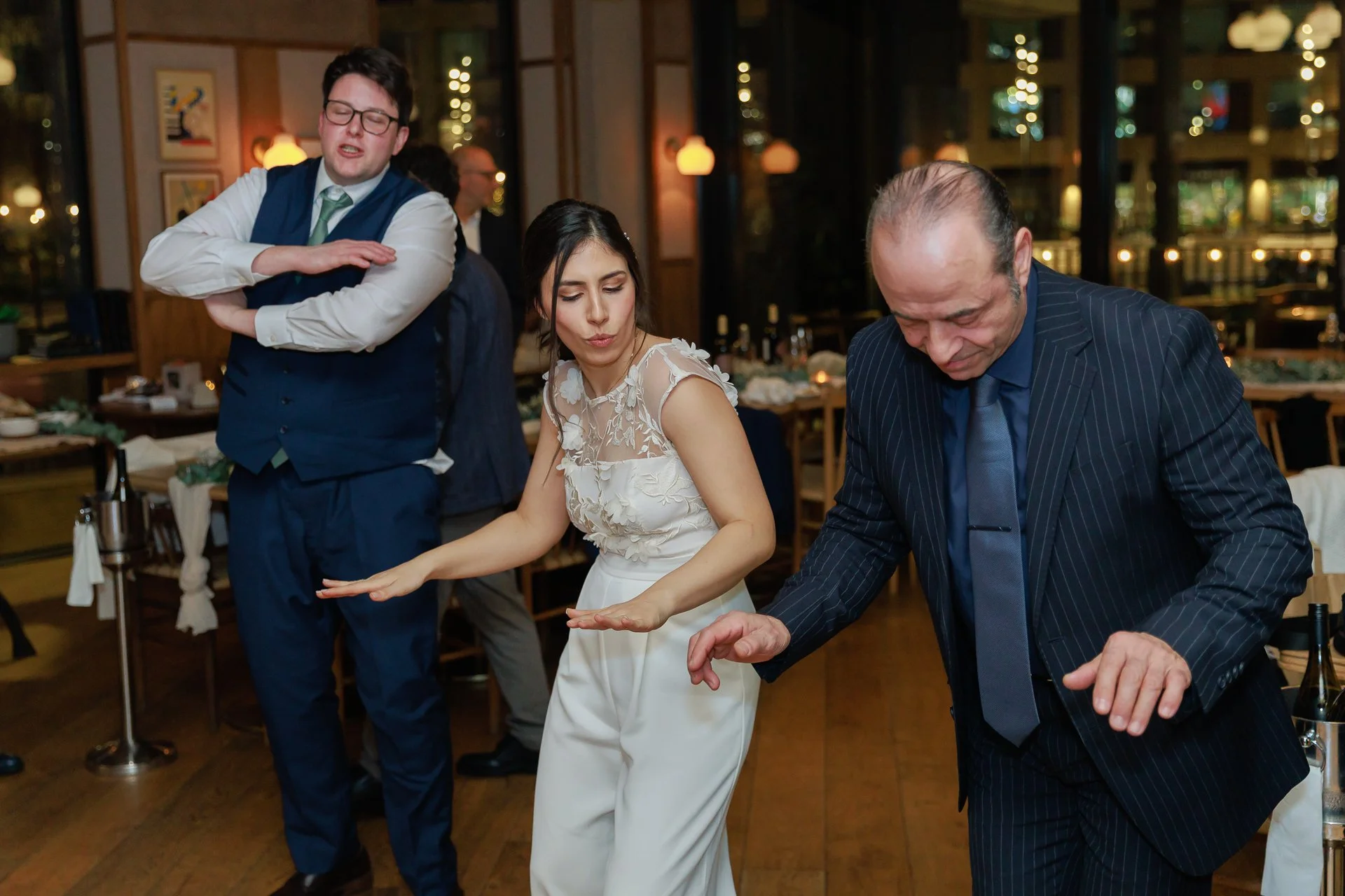 Three people dancing at a wedding reception. The woman in the middle wearing a white dress is dancing with an older man in a pinstripe suit, while a young man in a blue vest and glasses dances behind them.