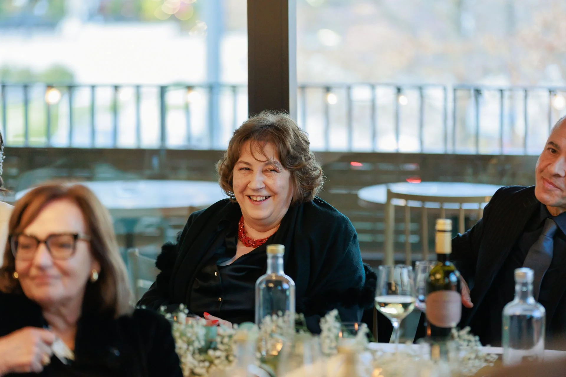 A woman with short, curly gray hair and a red necklace smiling at a dinner party with other guests, wine glasses, and a table decorated with flowers.