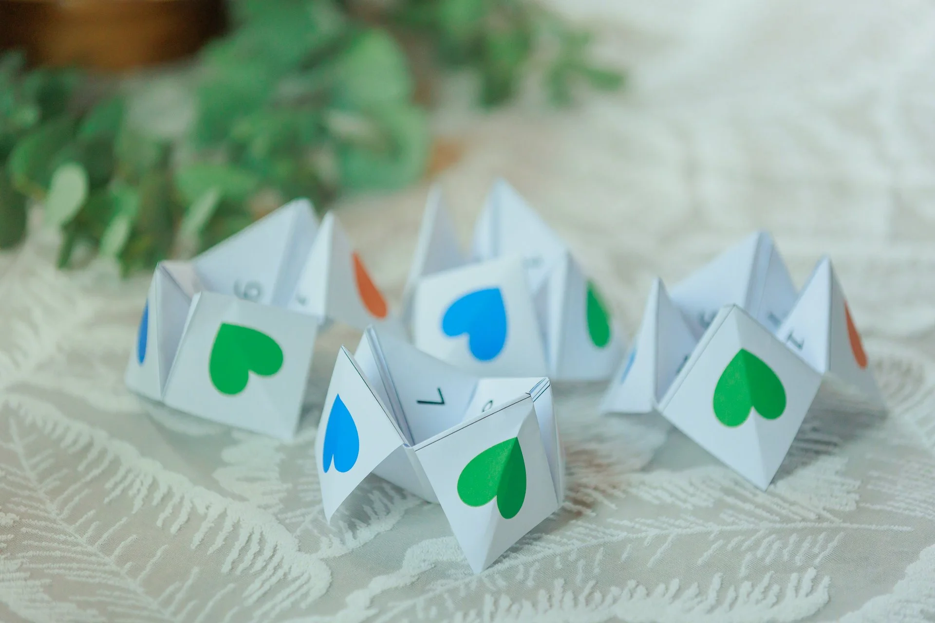 Colorful paper fortune teller with green, blue, and orange hearts on white background, resting on a lace tablecloth with green foliage in the background.