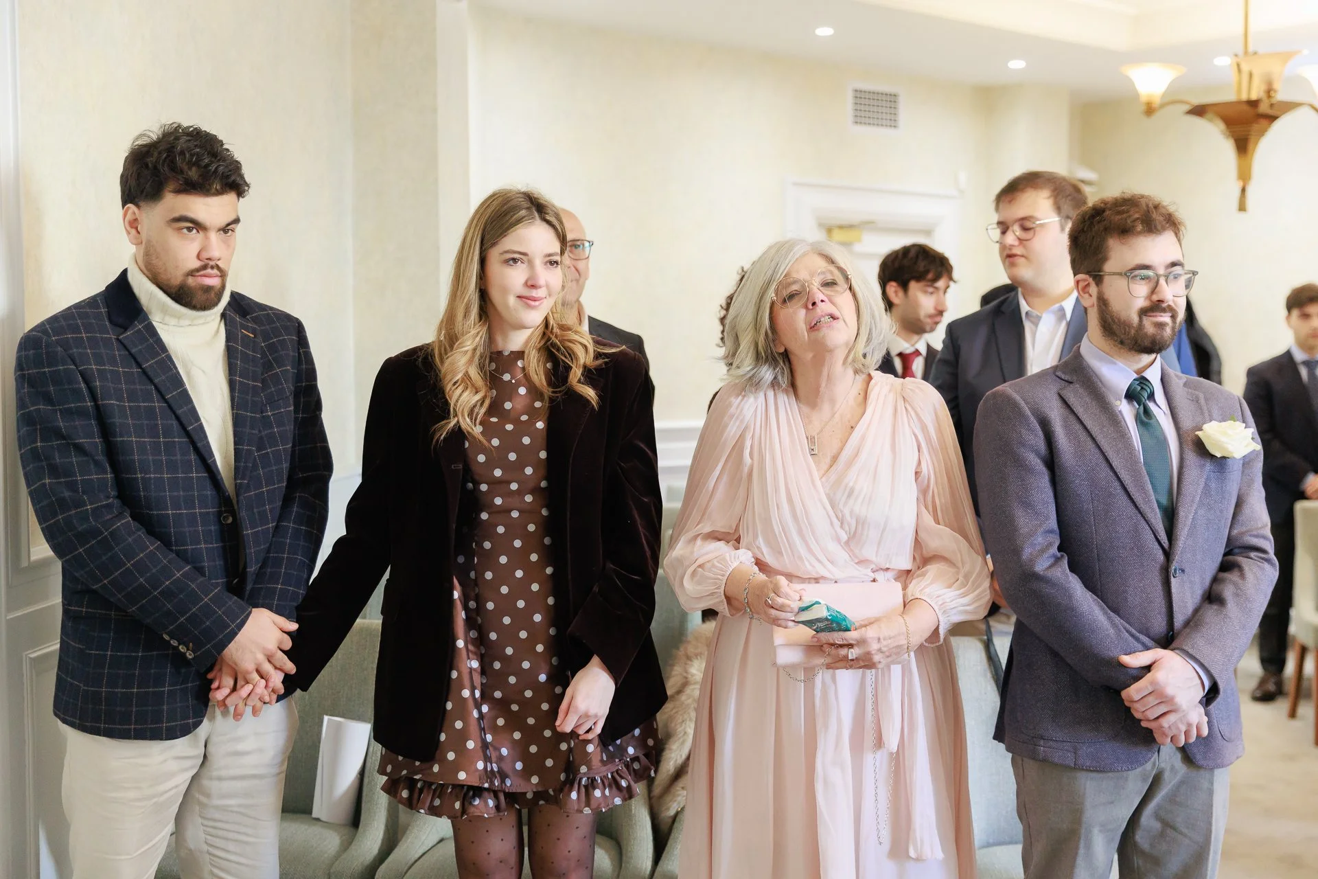Group of people attending a wedding ceremony, standing side by side, in a formal setting with cream-colored walls and elegant lighting.