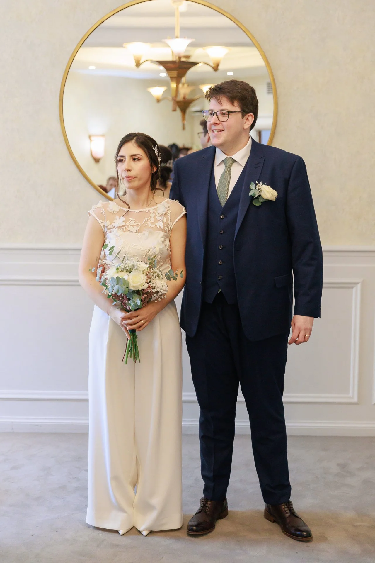 A bride and groom standing together at their wedding. The bride is holding a bouquet of white roses and greenery, wearing a lace top dress, and the groom is in a navy blue suit with a white shirt and a light-colored tie, smiling at the camera. They a