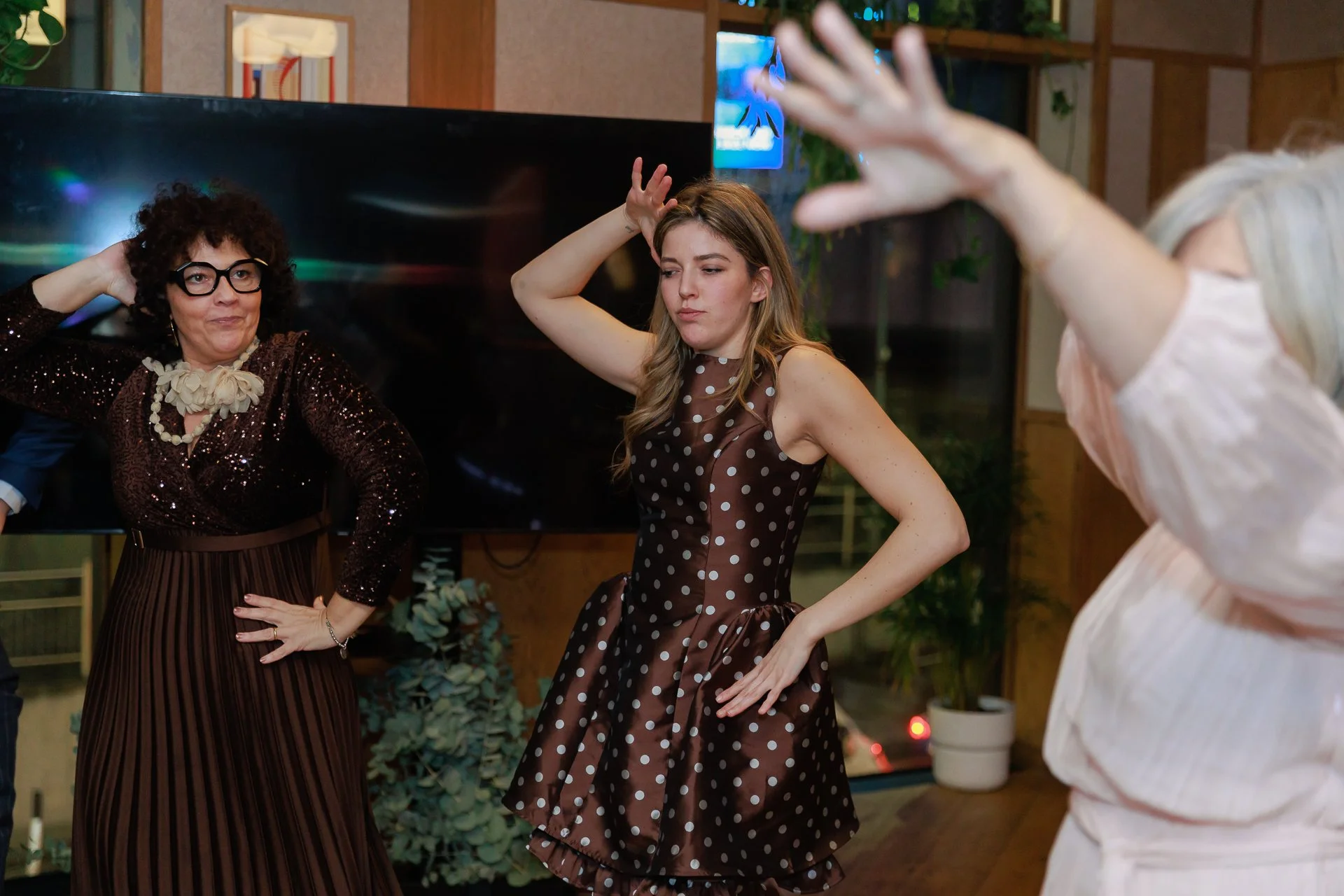 Three women dancing at an indoor social event with a TV screen and plants in the background.