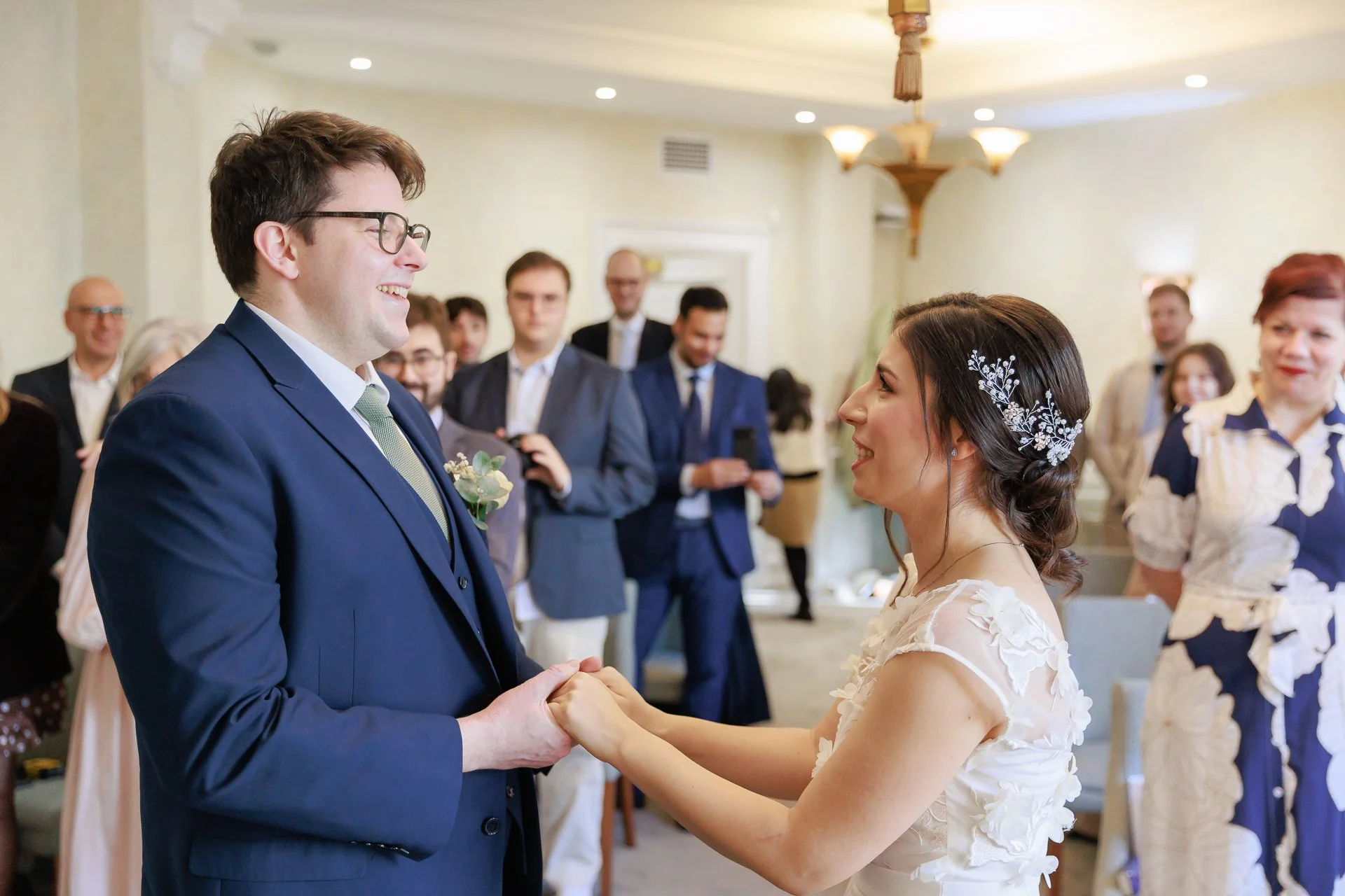 A bride and groom holding hands and smiling at each other during a wedding ceremony, surrounded by guests.