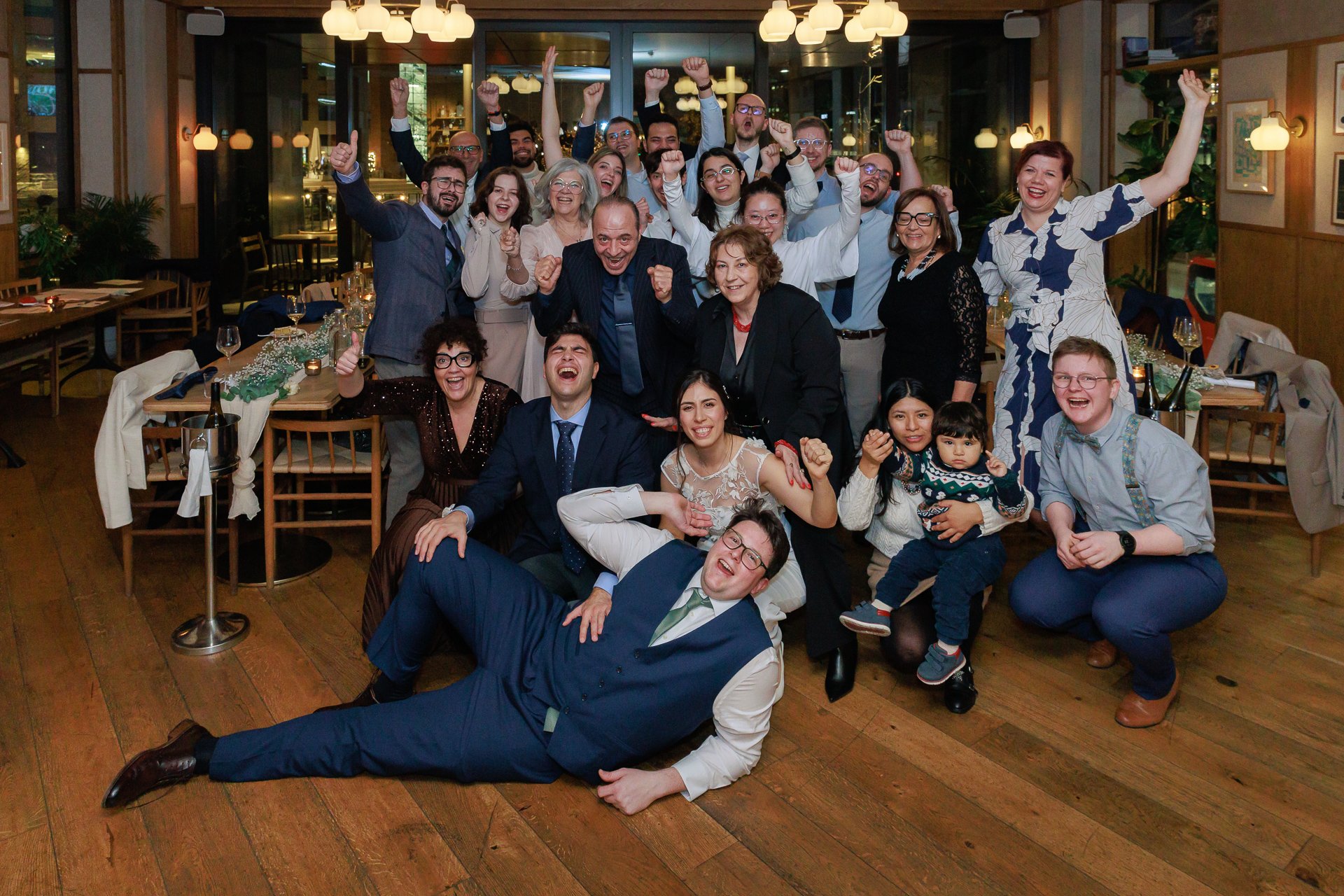 Group of people celebrating at a restaurant, smiling and making celebratory gestures, in a festive atmosphere with decorated tables and warm lighting.